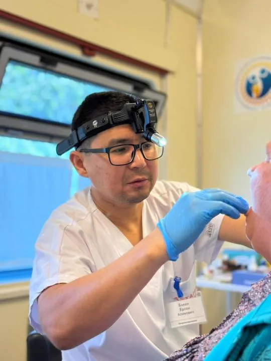 В центре — современный медицинский поезд с надписью «Саламатты Қазақстан». Рядом — местные жители, включая детей и пожилых, ожидают осмотра. Врачи в белых халатах встречают людей у входа.