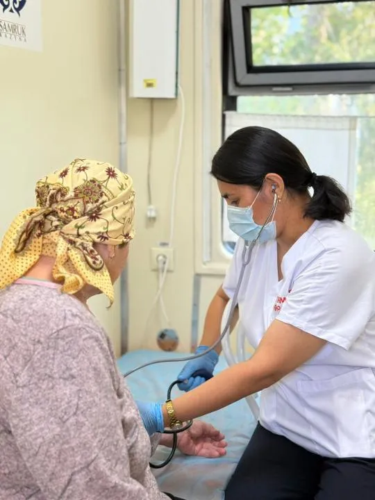 В центре — современный медицинский поезд с надписью «Саламатты Қазақстан». Рядом — местные жители, включая детей и пожилых, ожидают осмотра. Врачи в белых халатах встречают людей у входа.