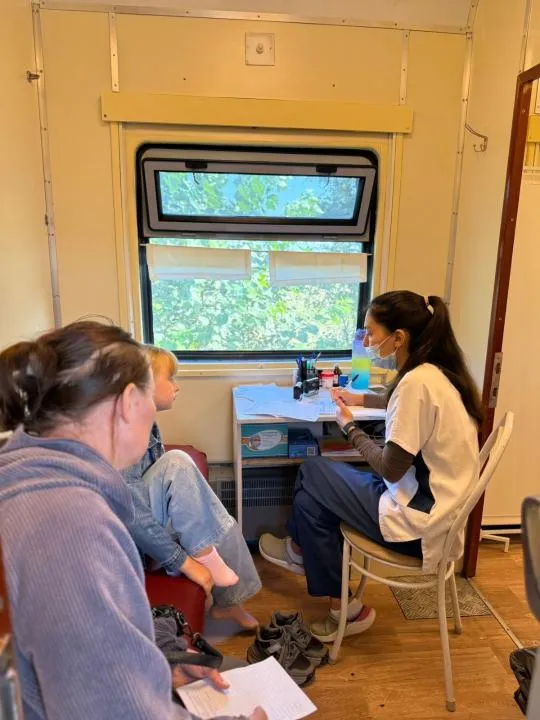 В центре — современный медицинский поезд с надписью «Саламатты Қазақстан». Рядом — местные жители, включая детей и пожилых, ожидают осмотра. Врачи в белых халатах встречают людей у входа.